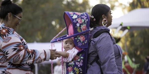 Indigenous woman wearing a baby in a carrier on her back while another woman holds the baby's hands