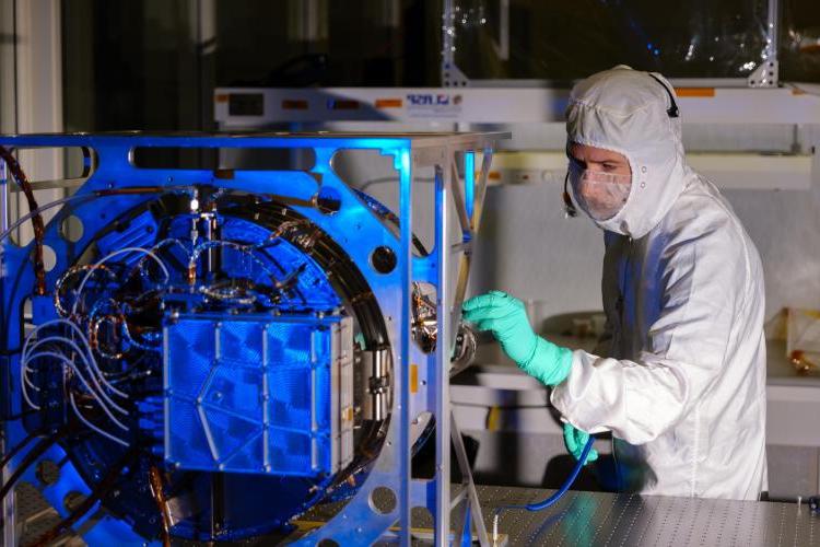 Engineer in protective gear inspects a metal space instrument with wires coming out of its back