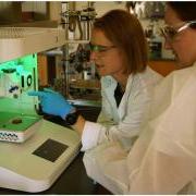 Two people in white lab coats, wearing safety goggles, work near lab equipment lit by a green light.