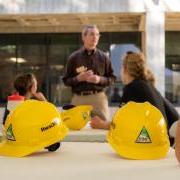 Dean Keith Molenaar stands before a group of people sitting at tables outdoors, with yellow construction hats on the tables.