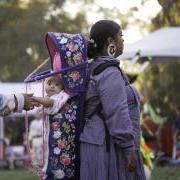 Indigenous woman wearing a baby in a carrier on her back while another woman holds the baby's hands